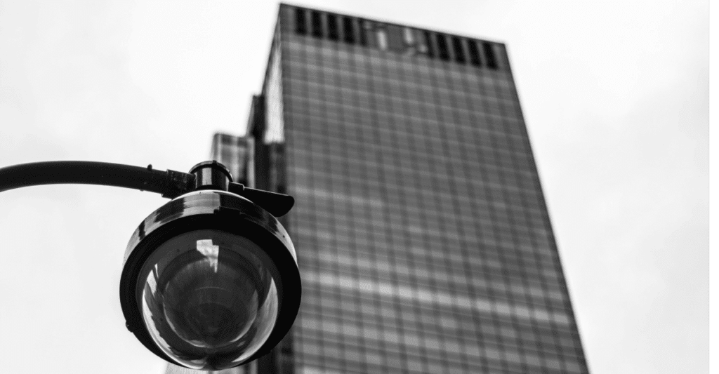 Here we have a black and white picture of a security camera in from of a skyscraper office block, connecting with the themes of big tech, surveillance, insight, and digital risk assessment.